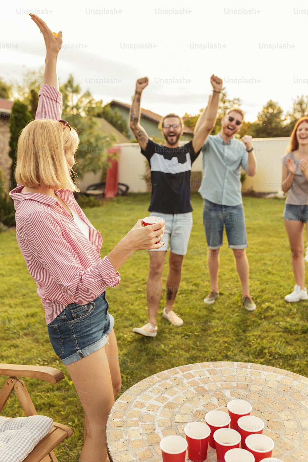 Gruppe fröhlicher junger Freunde, die Spaß haben, während sie Bierpong auf der Sommerparty im Hinterhof spielen und sich gegenseitig anfeuern