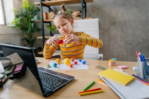 Kleines 8-jähriges Mädchen, das einen Laptop benutzt, um Online-Planeten des Sonnensystems zu studieren, sitzt am Schreibtisch im Raum. Junge Grundschülerin schaut sich online Astronomieunterricht an und macht ihre Hausaufgaben - modelliert Planetenmodelle aus Kinderton oder Plastilin.