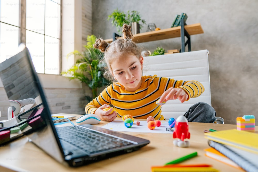 8 years old girl using laptop to study online planets of Solar system sitting on the desk in the room. Young female elementary school student watching astronomy lesson online and doing her homework - sculpts planets models from kids clay or plasticine.