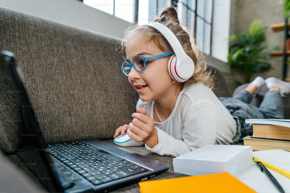 8 years old girl in the glasses studying online from home using laptop lying on the sofa in the room. Distance learning during coronavirus pandemic concept.