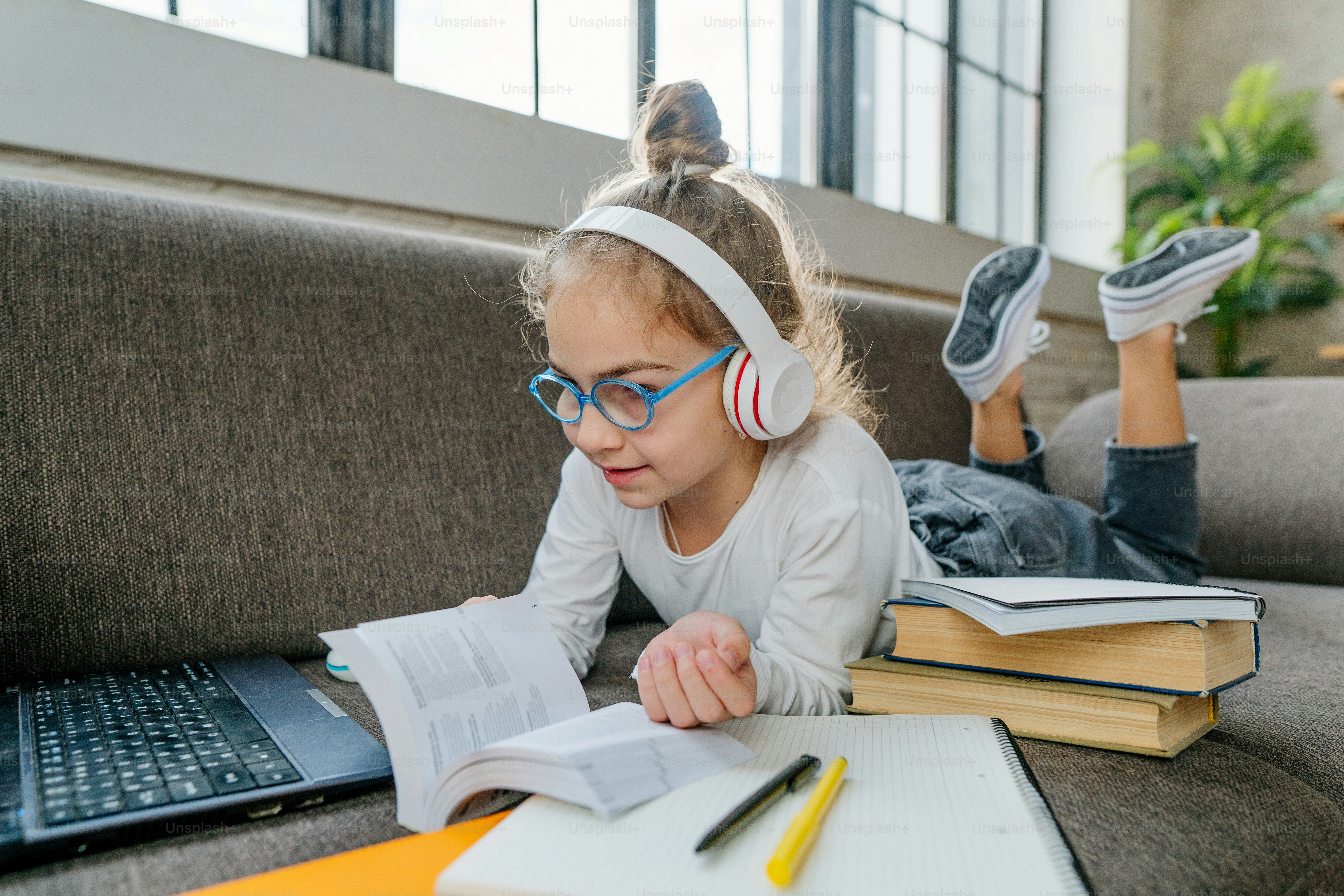 Fille de 8 ans dans les lunettes et les écouteurs lisant un livre étudiant en ligne à la maison à l’aide d’un ordinateur portable allongé sur le canapé de la chambre. Concept d’enseignement à distance pendant la pandémie de coronavirus.