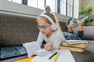 8 years old girl in the glasses and headphones reading book studying online from home using laptop lying on the sofa in the room. Distance learning during coronavirus pandemic concept.