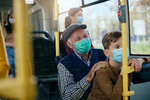Homem idoso feliz e seu neto viajando de ônibus público e usando máscaras faciais de proteção devido à pandemia de coronavírus.