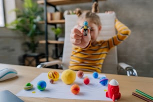 8 years old girl study Solar system sitting at the desk in the room. Female elementary school student doing her homework - sculpts planets models from kids clay or plasticine. Selective focus on the astronaut.