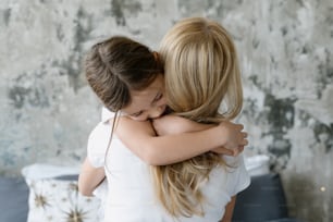 Charming and smiling little daughter hug her loving mother while they spending free time together at home. Woman sits with her back to the camera and cuddle to kid girl