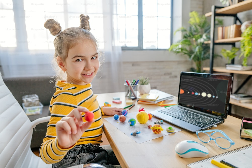 Little 8 years old girl using laptop to study online planets of Solar system sitting at the desk in the room. Young female elementary school student watching astronomy lesson online and doing her homework - sculpts planets models from kids clay or plasticine.