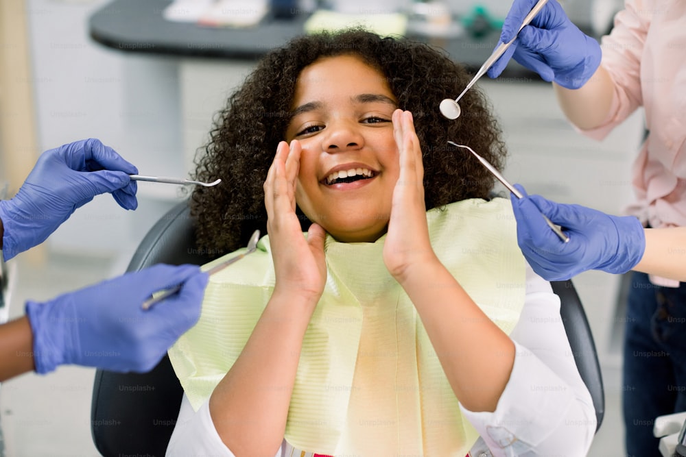 Healthy teeth, caries prevention and pediatric dentistry. Close up of scared funny little mixed raced a girl, looking at camera and screaming and hands of two dentists in gloves with tools.