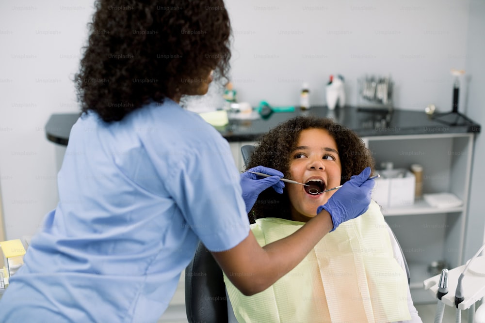 Close up of pretty a mixed raced teen girl in dentist chair, with mouth open, having dental examination. Back view of female African dentist in gloves, holding dental tools.
