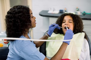 치과 진료실에서 치과 치료를 받는 작은 아프리카계 미국인 소녀 환자의 클로즈업 보기. 드릴과 치과 거울을 사용하여 치아 우식증 치료를 만드는 여성 흑인 치과 의사.