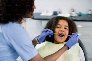 Close up top view of pretty kid, african american curly girl in dentist chair, with mouth open, having dental examination. Back view of female African dentist in gloves, holding tools.