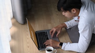 Jovem asiático trabalhando on-line em laptop e deitado no chão em casa.