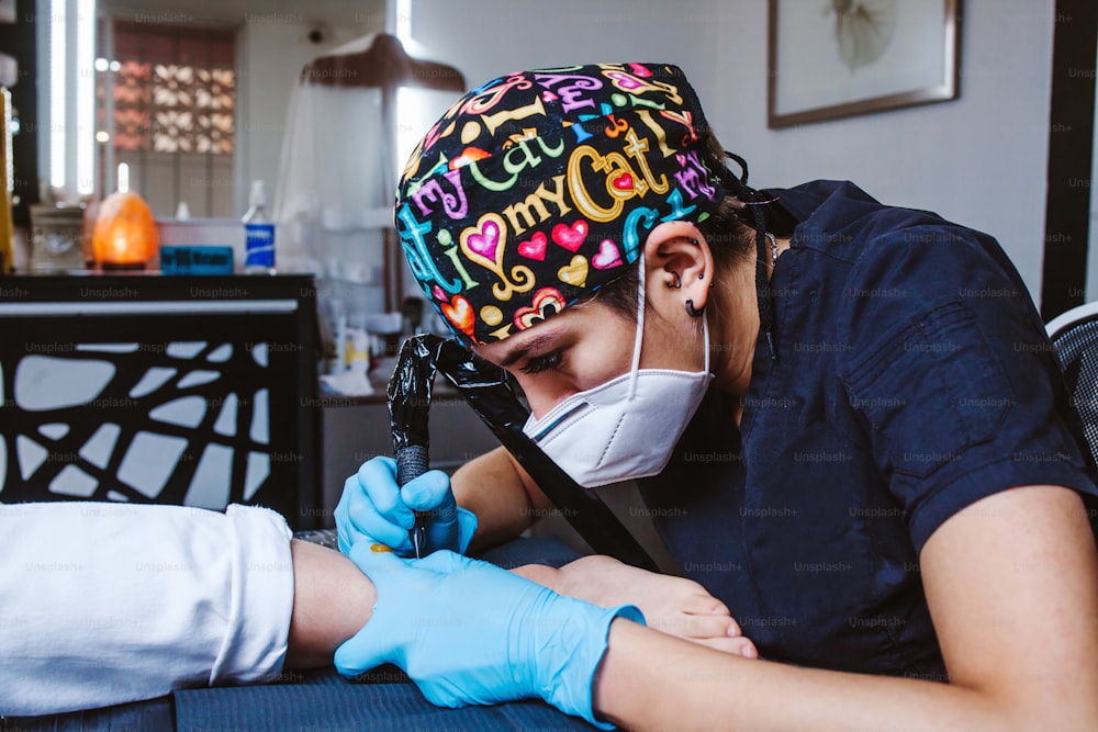 ラテン系の女性タトゥーアーティストが、メキシコシティで青い滅菌手袋を着用し、ペイントでタトゥーを入れるプロセスを実演しています