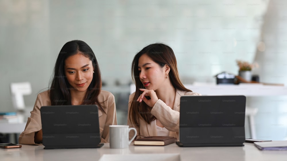 Deux femmes d’affaires confiantes partageant des idées et travaillant ensemble avec une tablette dans un bureau moderne.