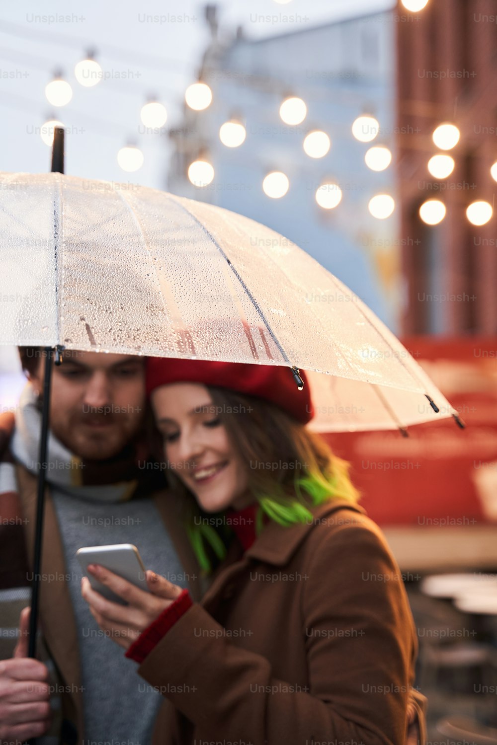 Vertical view portrait of the enamored stylish couple standing at the empty autumn street and watching their photos at the smartphone while walking. Focus at the umbrella with drops