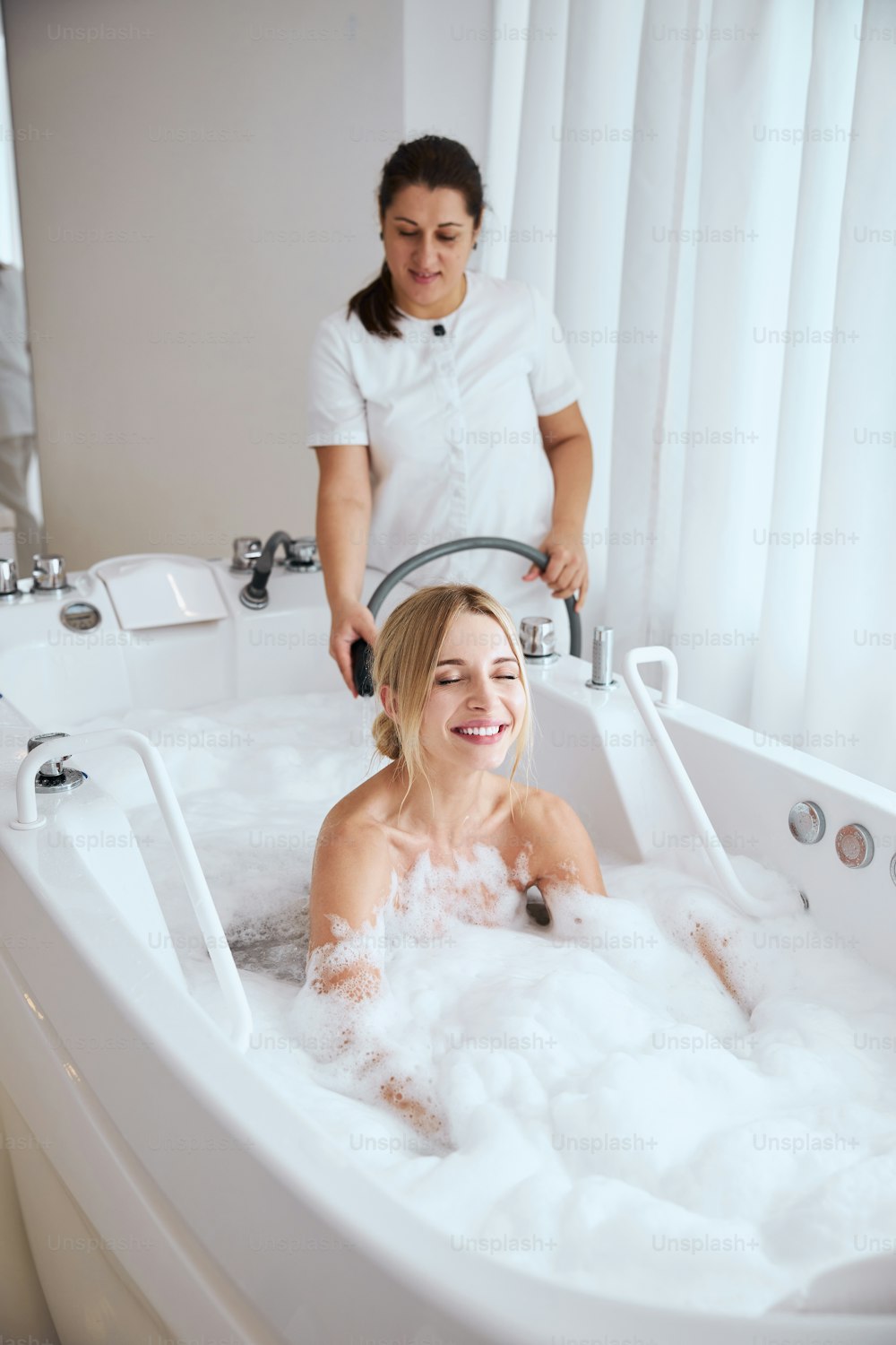Waist up portrait of female enjoying in the hydro massage bathtub and professional spa salon worker using a hose during the underwater massage