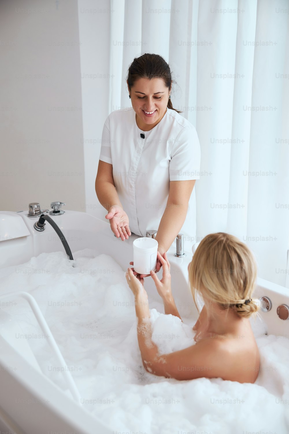 Back view portrait of elegant pretty female sitting in hydro massage bath tube and professional female worker demonstrating jar with cream for patient