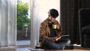 Retrato de homem casual freelancer e usando tablet digital e beber café enquanto sentado no chão perto da janela.