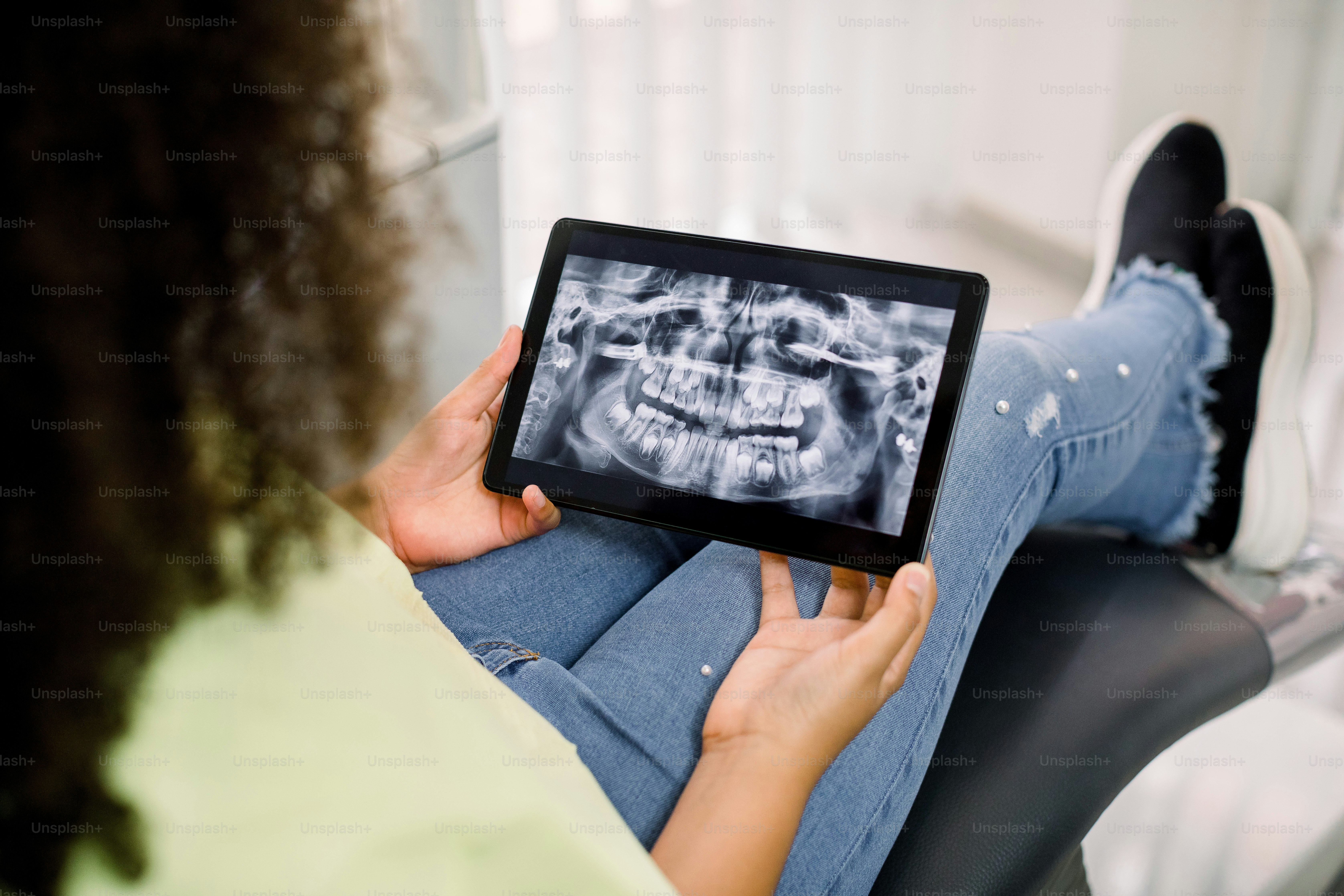 Rückwärtige Nahaufnahme abgeschnittene Ansicht eines kleinen geduldigen, lockigen afroamerikanischen Teenagermädchens, das in einer modernen Klinik auf dem Zahnarztstuhl sitzt und einen digitalen Tablet-PC mit Röntgenpanoramabild von Zähnen und Kiefern hält.
