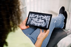 Vista ritagliata ravvicinata posteriore di un piccolo paziente, ragazza adolescente afroamericana ricciuta, seduta sulla poltrona odontoiatrica in una clinica moderna, tenendo in mano un tablet pc digitale con un'immagine panoramica a raggi X di denti e mascelle.