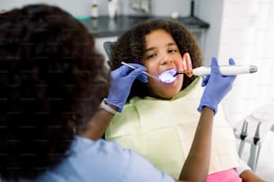 小児歯科医院で少し混血の女子高生に歯科充填手順を行うアフリカの女性歯科医。充填重合に歯科用紫外線硬化光を使用する医師。
