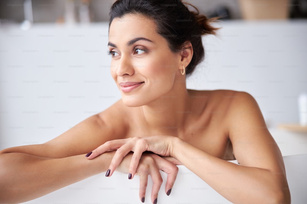 Woman is lying in the bathroom and looking sideways, she holding her hands on the edge of the tub. Stock photo