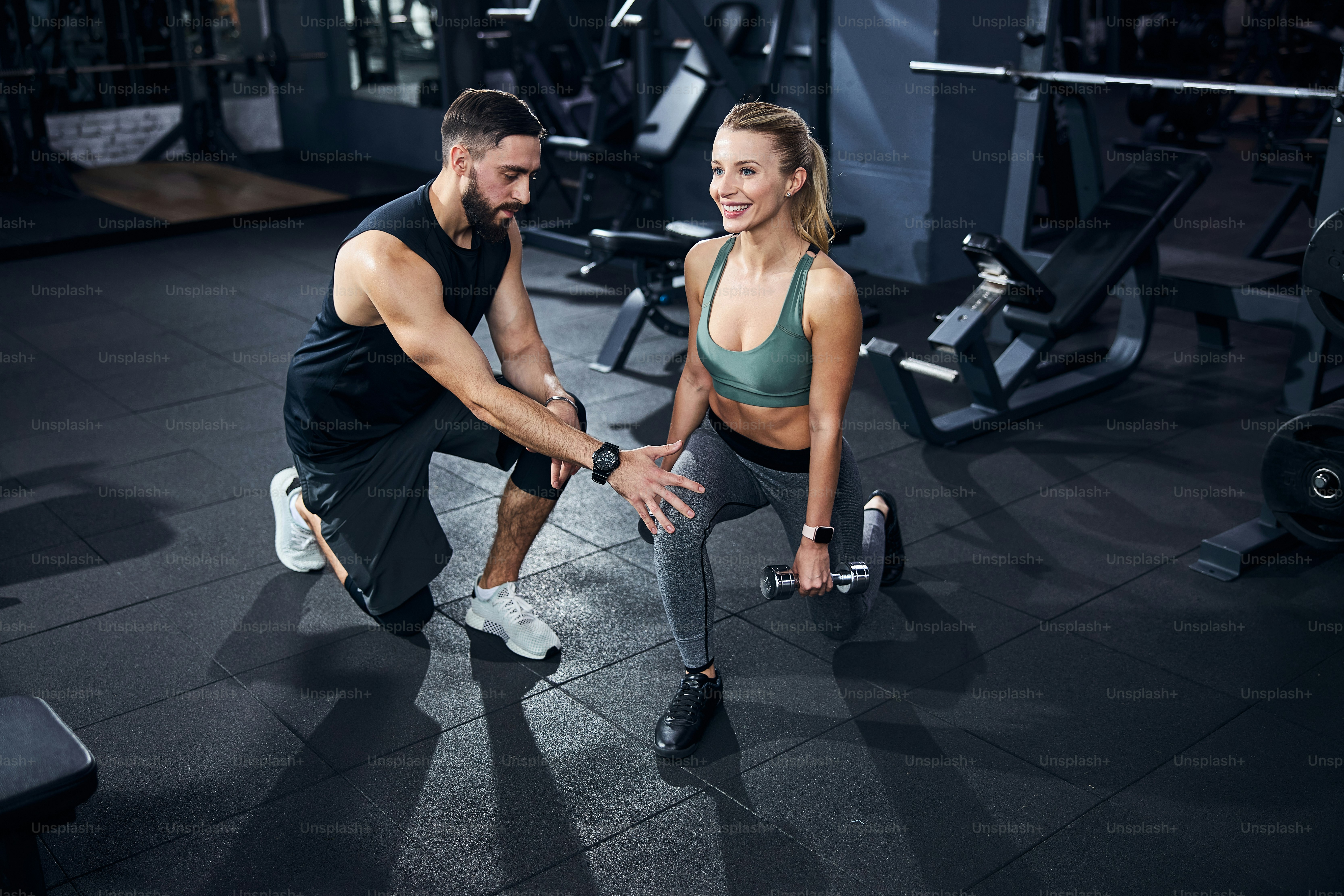 Man wearing dark clothes fixing the leg position of a female athlete before the start of dumbbell activity