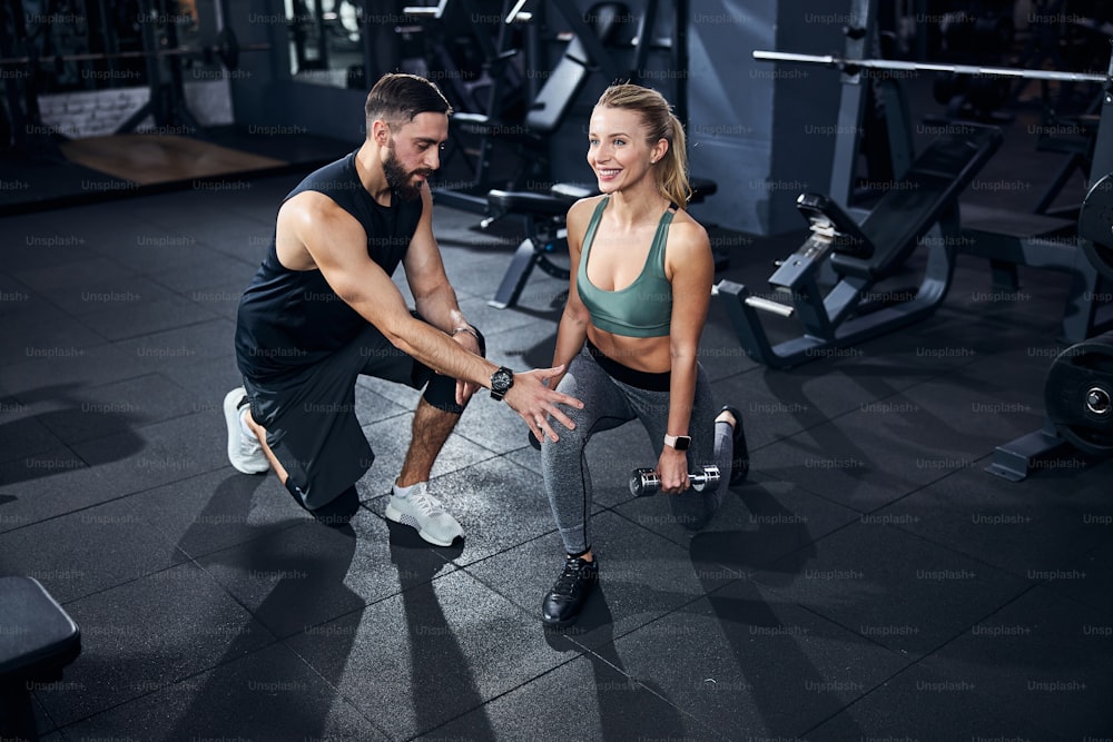 Man wearing dark clothes fixing the leg position of a female athlete before the start of dumbbell activity