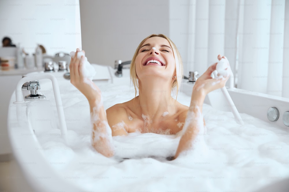 Close up portrait of funny Caucasian female having fun with foam while lying on the luxury bathtub in room indoors