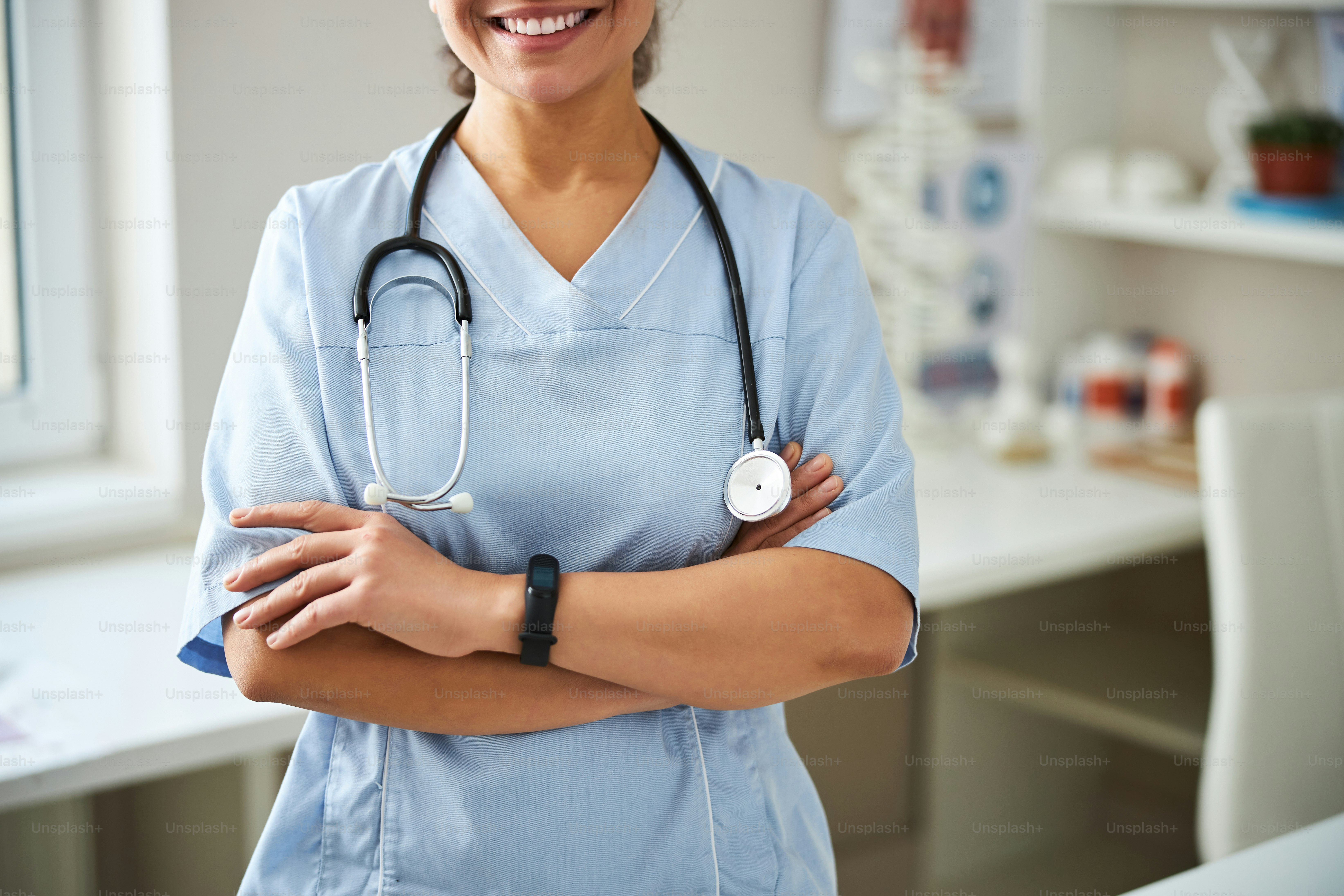 Cropped photo of a lady standing in a clinic room and smiling while folding her arms on her breast
