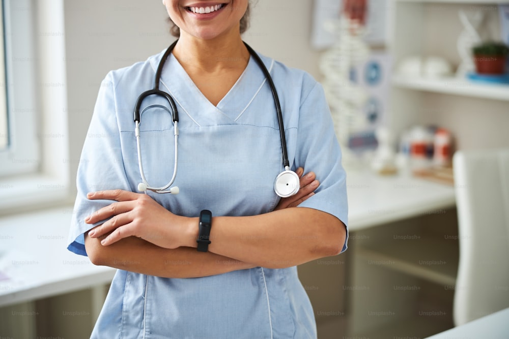 Foto recortada de una señora de pie en una habitación de clínica y sonriendo mientras cruza los brazos sobre su pecho