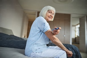 Adulta jubilada feliz sentada en una cama y haciéndose una autoinyección en el brazo por debajo del hombro