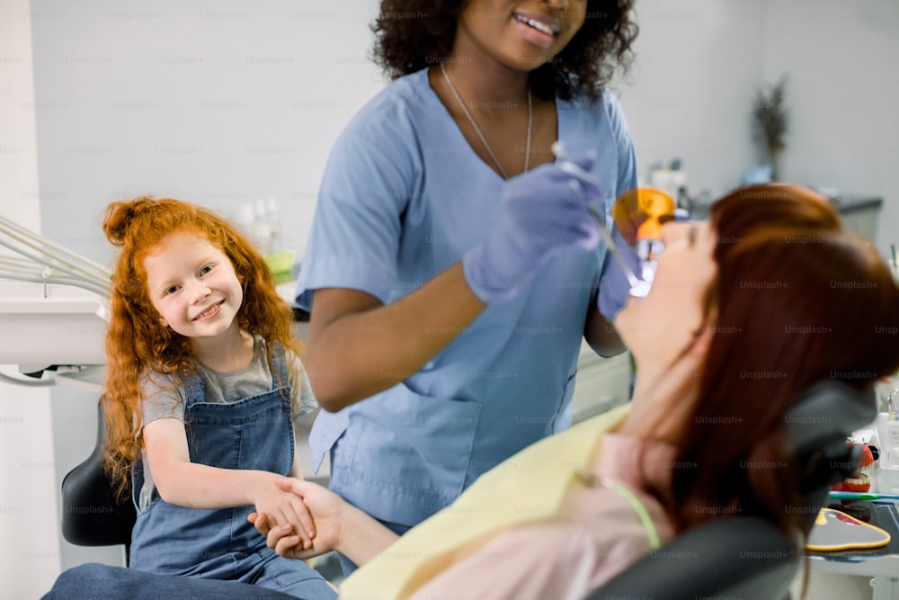 Young little red haired curly girl supporting her mom at dentist's office during checkup. Female smiling African pediatric dentist in blue uniform and gloves makes teeth filling with UV lamp.