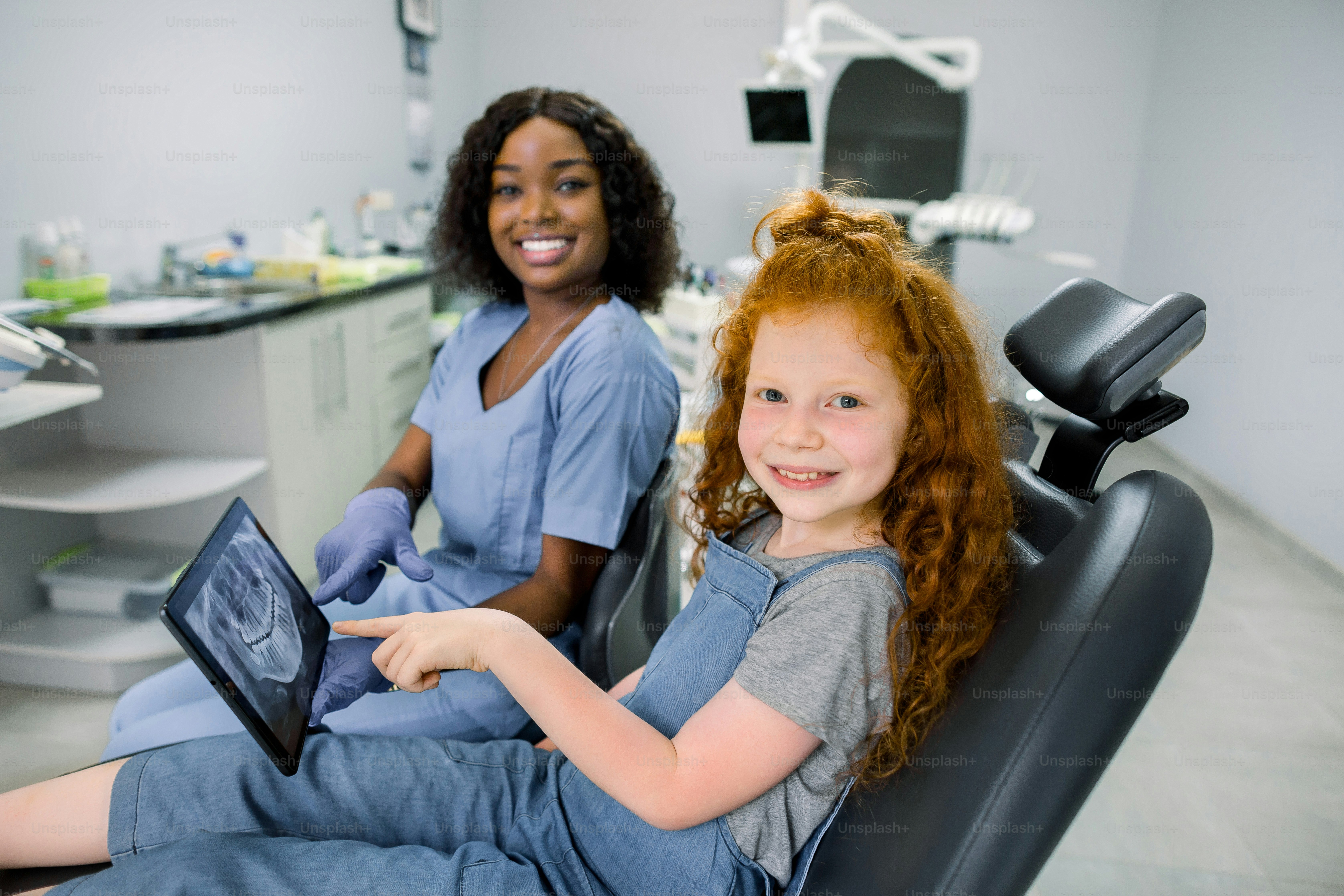 Niño en el consultorio del dentista. Pequeña niña paciente de cabello rojo, sentada en la silla de odontología, mirando a la cámara y señalando en la tableta con el escaneo panorámico de los dientes junto con su dentista africano sonriente.