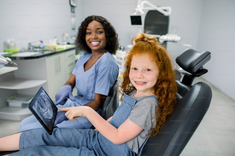 Child at dentist's office. Little patient red haired girl, sitting in dentistry chair, looking at camera and pointing on tablet with teeth panoramic scan together with her smiling african dentist.
