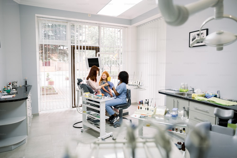 Modern dentist's office. Mother and little daughter having a regular check up and talking with their dentist, young African woman. Oral care, caries prevention.