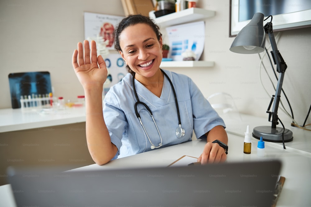 Joyful physician raising her arm and giving a wave to a screen during an online video meeting