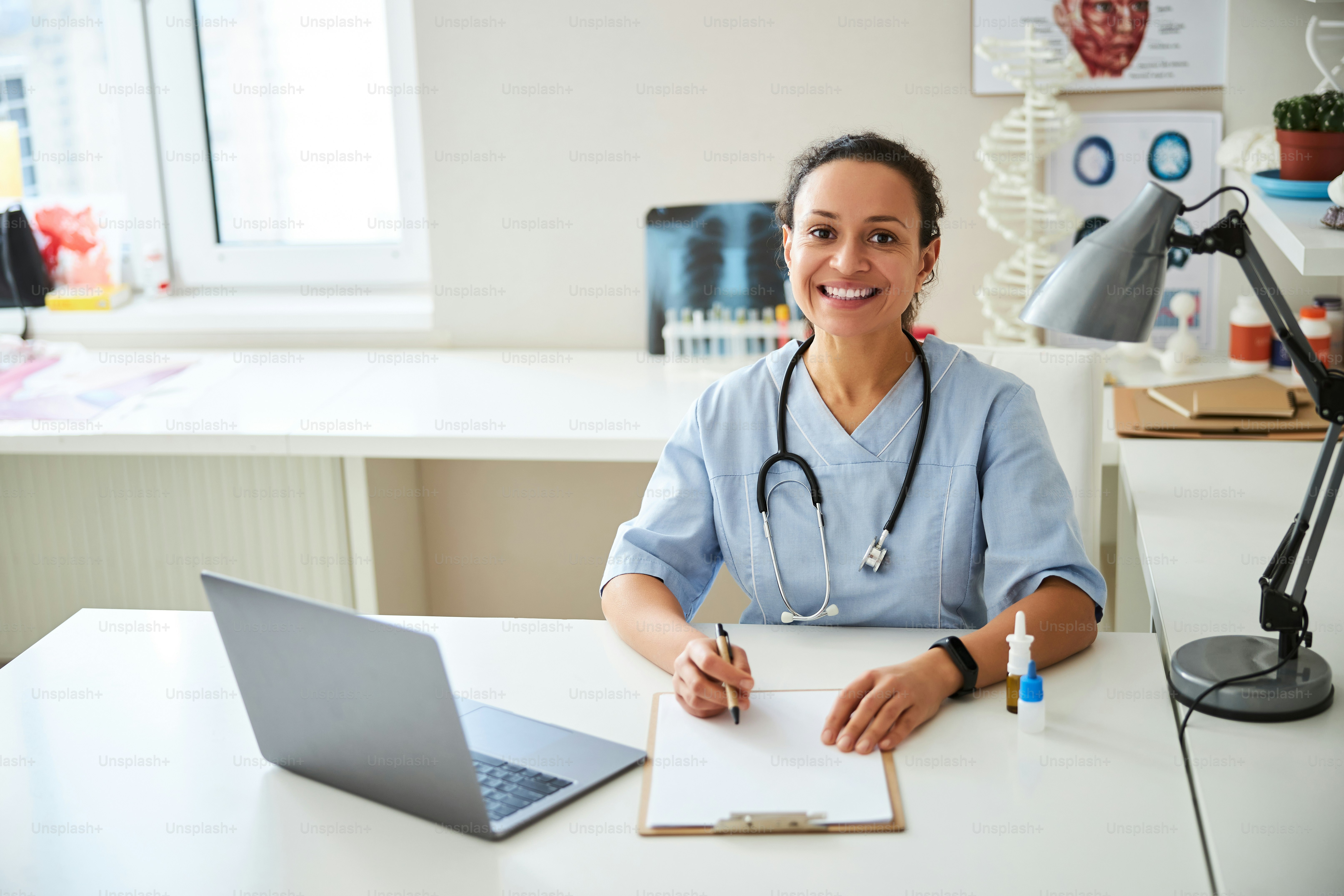 Doctor smiling at the desk in front of the laptop with a stethoscope  hanging from her neck photo – Laptop Image on Unsplash, image size:3000x2000