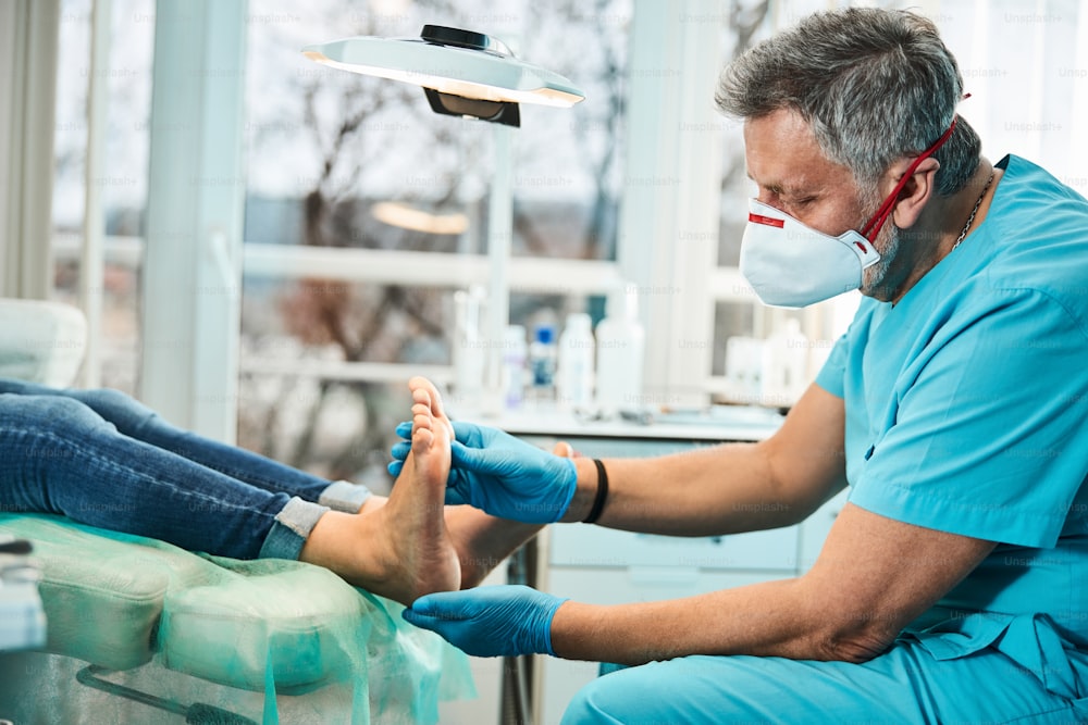 Retrato de vista lateral del podólogo barbudo adulto barbudo que examina los pies y los dedos de los pies mientras el paciente está sentado en la silla médica en la clínica de belleza