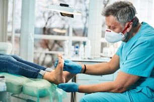 Side view portrait of adult bearded man chiropodist examining feet and toes while patient sitting on the medical chair in beauty clinic