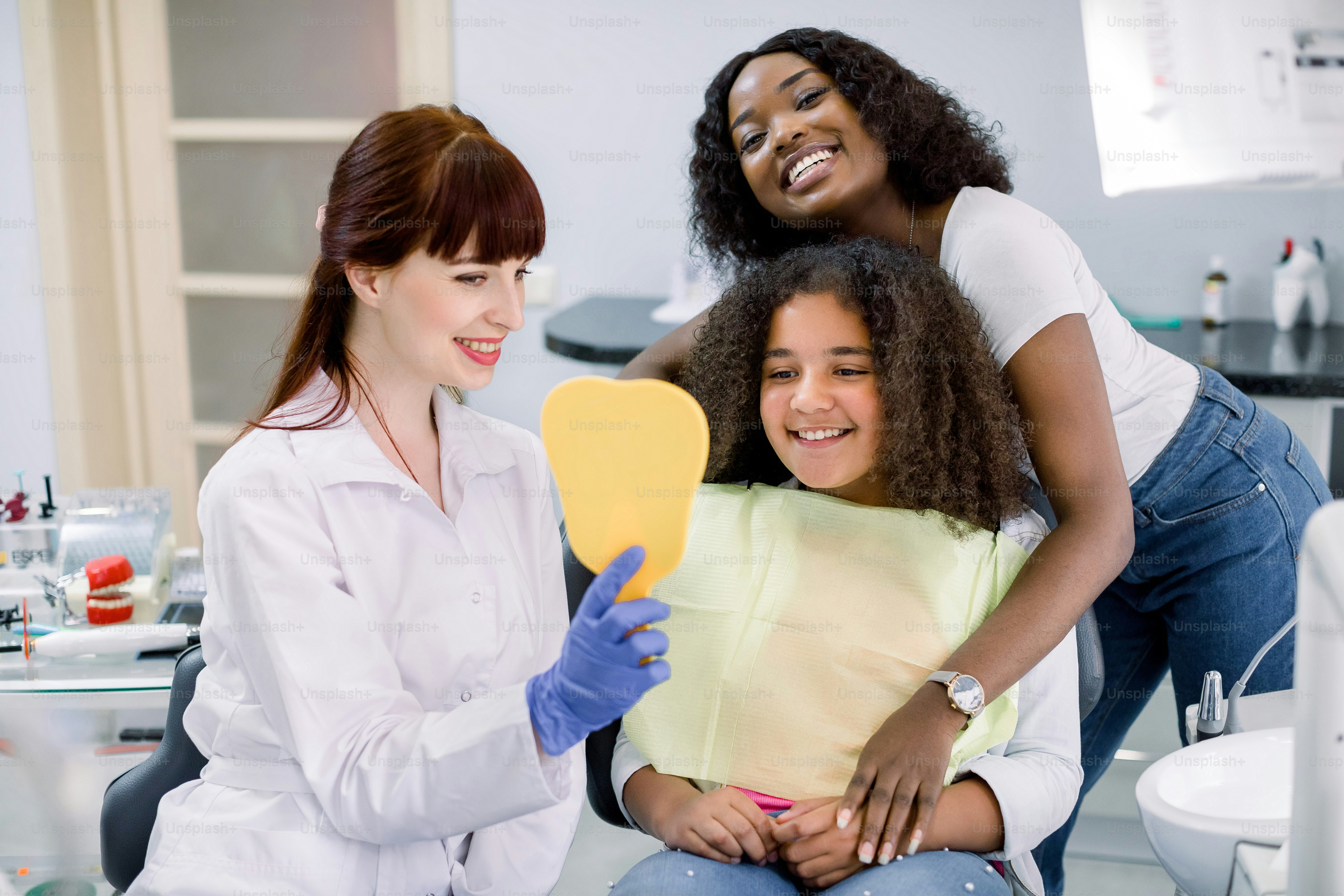 Cute African schoolgirl visiting dentist's office with her mom, seating in dentist chair and looking at the mirror on her beauty smile, while happy woman dentist shows her result of treatment.