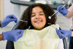 Charming little African American girl afro hair, sitting in dental chair, smiling and looking at camera during medical treatment at modern dental clinic. Hands of two dentists with dental tools