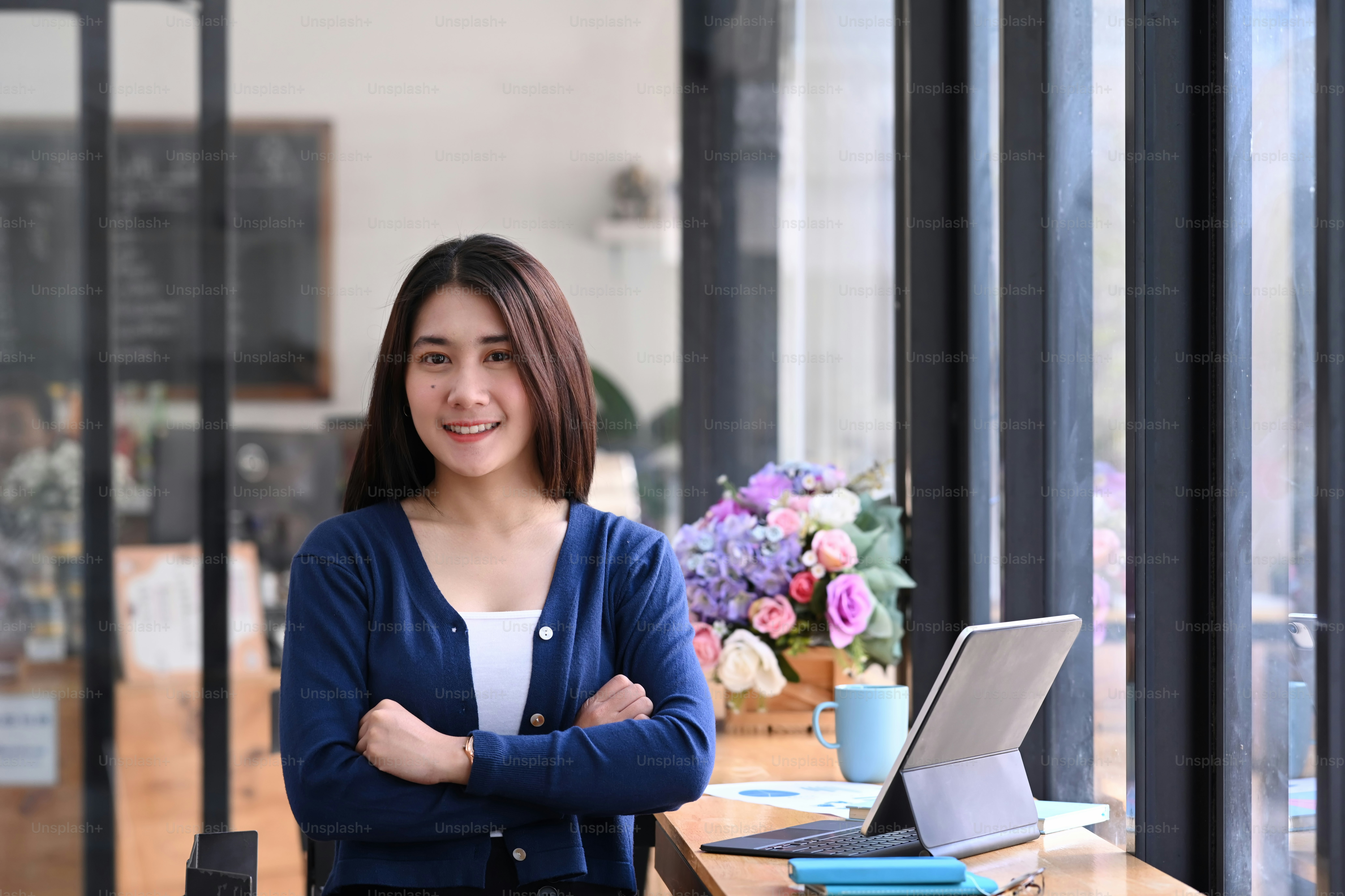 Une jeune femme joyeuse, les bras croisés, debout dans un bureau moderne et souriant à la caméra.