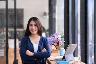A cheerful young woman with arms crossed standing in modern office and smiling to camera.