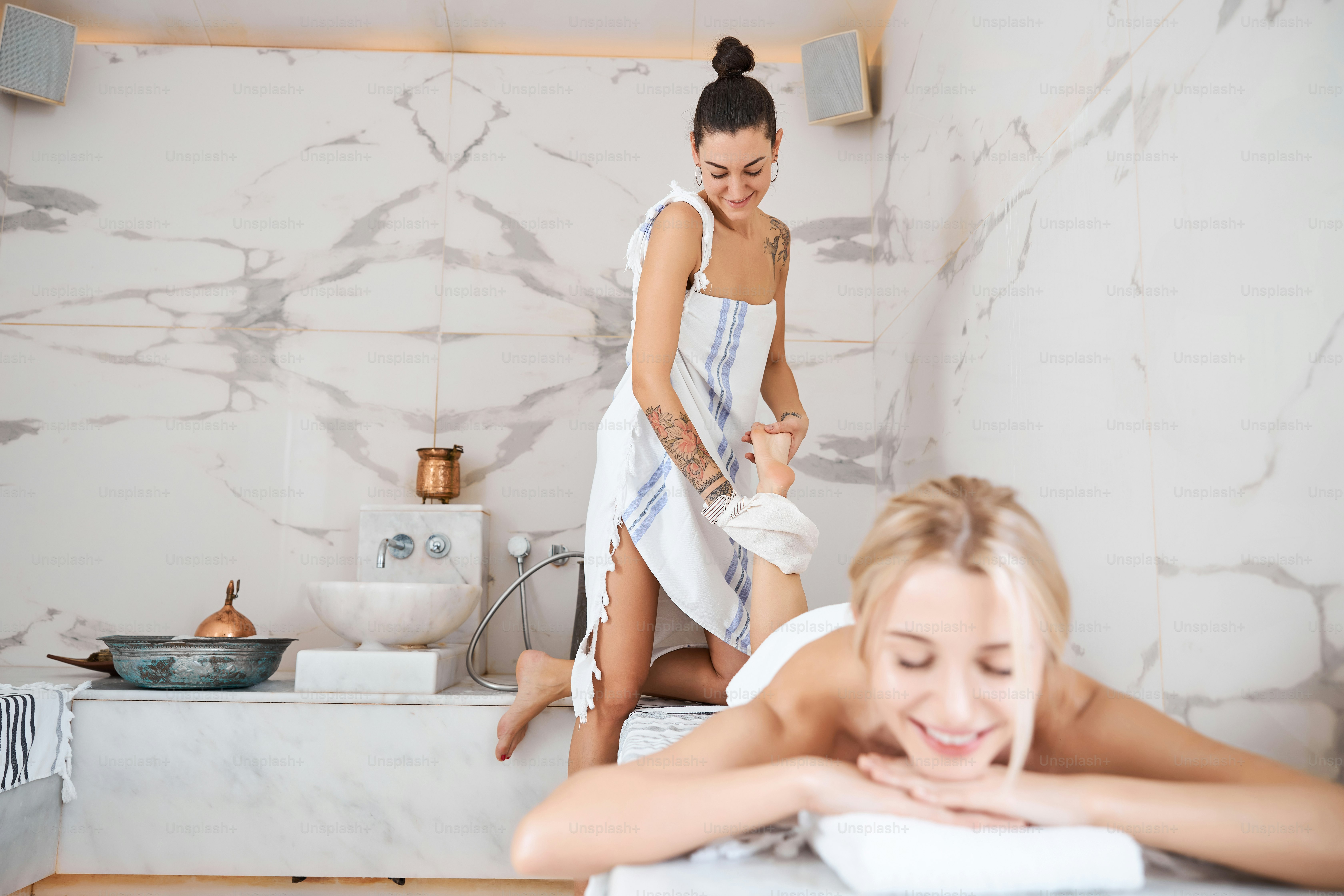 Potret panjang penuh pekerja hammam rambut gelap dengan handuk peshtemal melakukan prosedur spa untuk klien wanita cantik di dalam ruangan