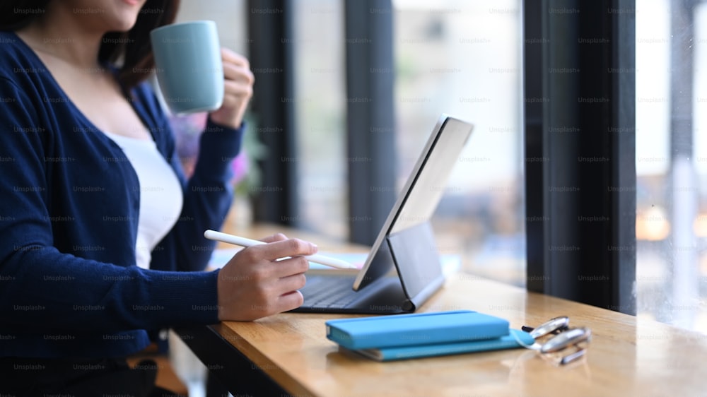 Ausschnittaufnahme einer jungen Frau, die Kaffee trinkt und mit einem Tablet-Computer arbeitet, während sie an ihrem Arbeitsplatz sitzt.