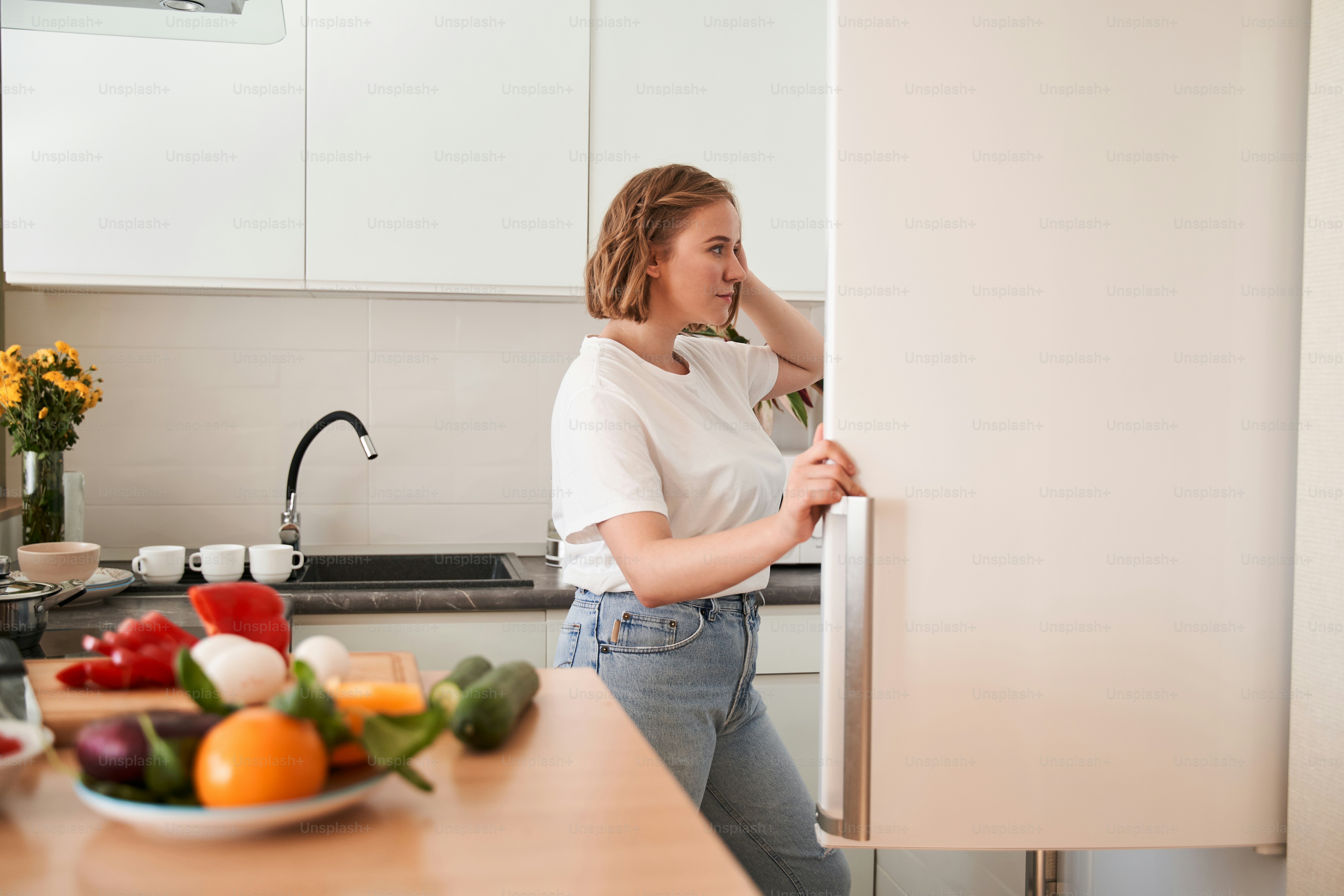 Vista lateral de la linda mujer caucásica abriendo su refrigerador mientras busca ingredientes para su plato culinario mientras pasa tiempo en la cocina
