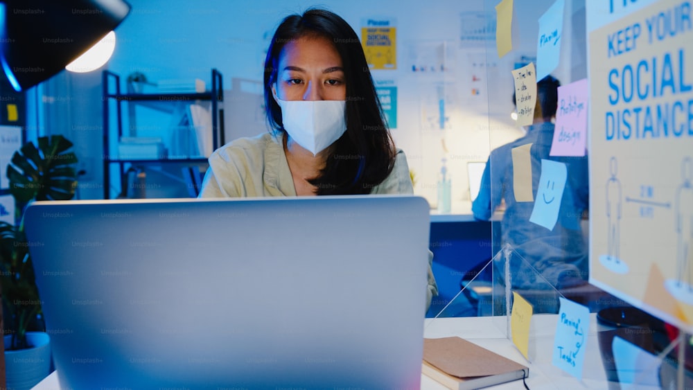 オフィスの夜に職場でラップトップを使用しながら、ウイルス予防のための新しい通常の状況で社会的距離を保つために医療用フェイスマスクを着用している幸せなアジアのビジネスウーマン。コロナ後の生活と仕事。
