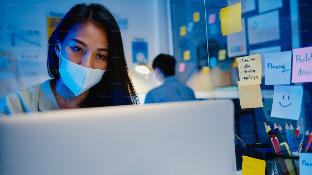 Happy Asia businesswoman wearing medical face mask for social distancing in new normal situation for virus prevention while using laptop back at work in office night. Life and work after coronavirus.