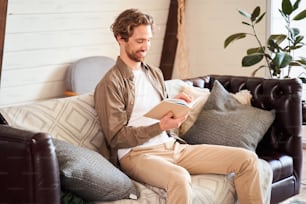 Portrait of nice cheerful stylish young man sitting on the sofa, holding in hands book and reading with pleasure emotions in the light modern interior