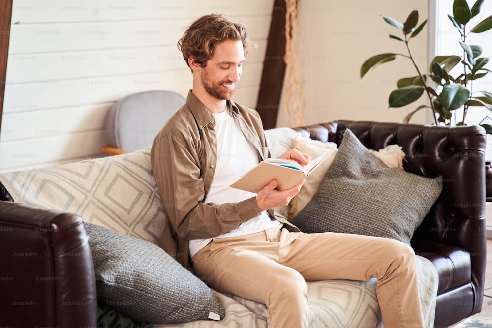 Portrait of nice cheerful stylish young man sitting on the sofa, holding in hands book and reading with pleasure emotions in the light modern interior